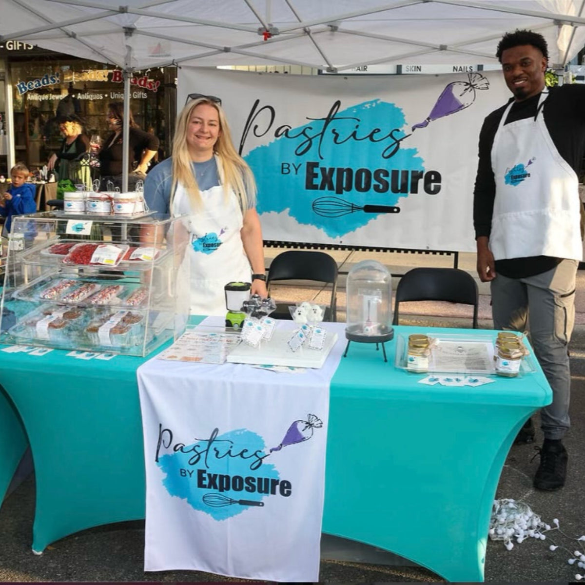 Two people behind a table with 'Pastries by Exposure' branding at an outdoor event.
