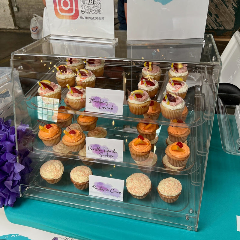 Clear display case with cupcakes on a table with 'Exposure' branding.