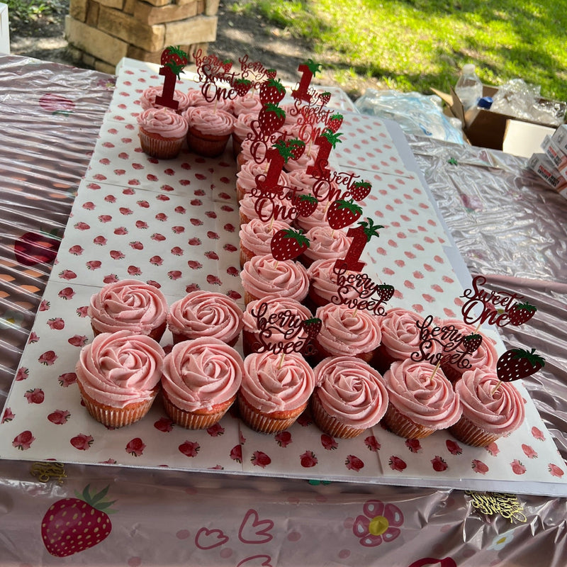 Pink cupcakes with 'Sweet One' decorations on a polka dot tablecloth outdoors.