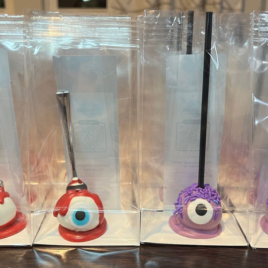Two novelty eye-shaped spoons in clear packaging on a wooden surface.
